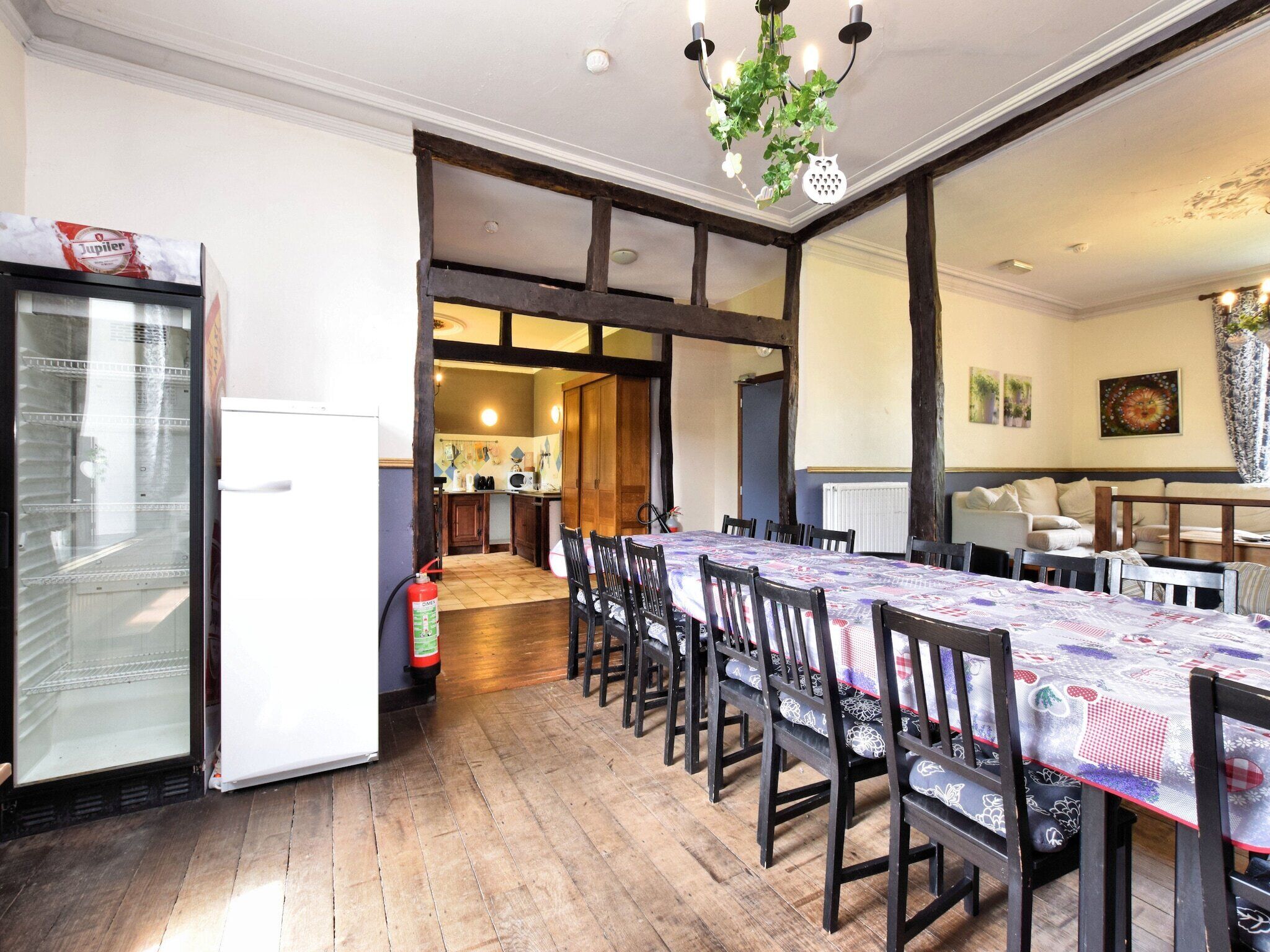 A dining area with a long table, chairs, and a chandelier. There is a kitchen visible in the background.