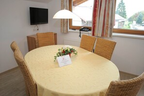 A dining area with a round table, wicker chairs, a floral centerpiece, a welcome card, a flat-screen TV mounted on the wall, and a window with curtains.