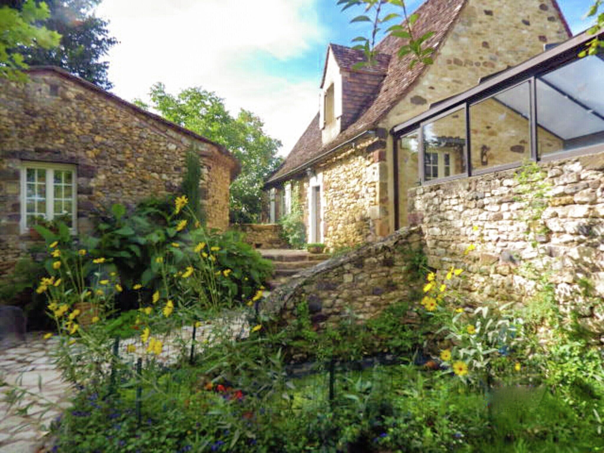 Un bâtiment en pierre avec un toit de chaume, une véranda en verre et un jardin où poussent diverses plantes et fleurs.