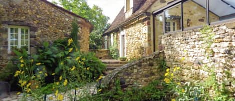 A stone building with a thatched roof, a glass conservatory, and a garden with various plants and flowers.