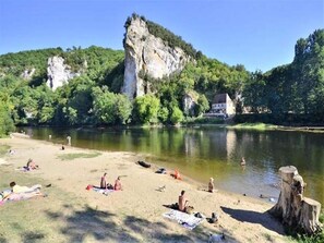 A riverside beach with people sunbathing and swimming, surrounded by greenery and a large rock formation. - Grives Poolside Family Escape (Belvès)