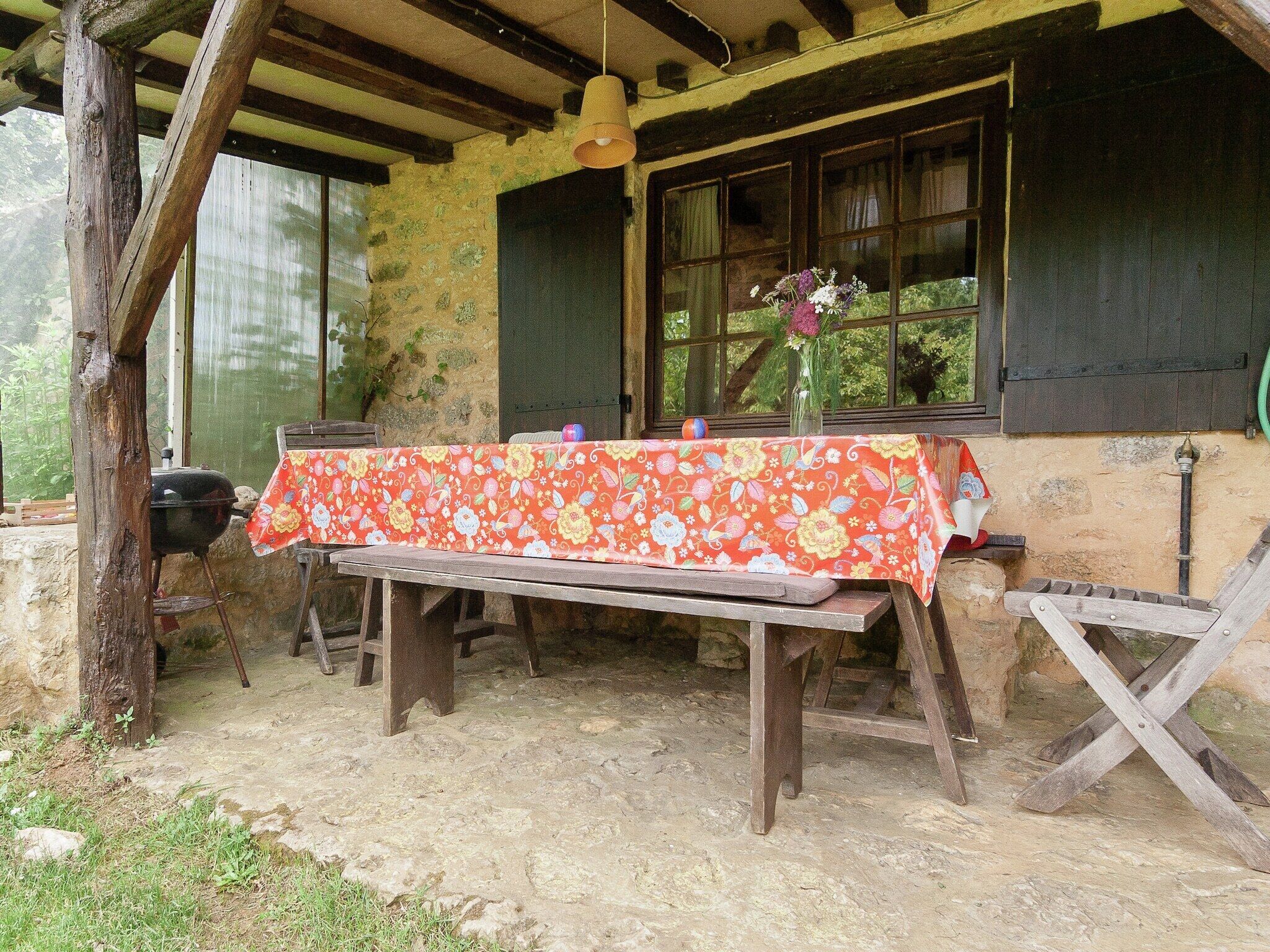 Une table en bois placée à l’extérieur, recouverte d’une nappe à motifs floraux, avec une suspension au-dessus et une fenêtre agrémentée de rideaux.