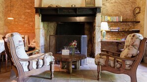 A rustic room with a fireplace, two upholstered chairs, a round wooden table, and a bookshelf filled with books. - Holiday Home in Dordogne near Lascaux Cave (Les Eyzies-De-Tayac-Sireuil)