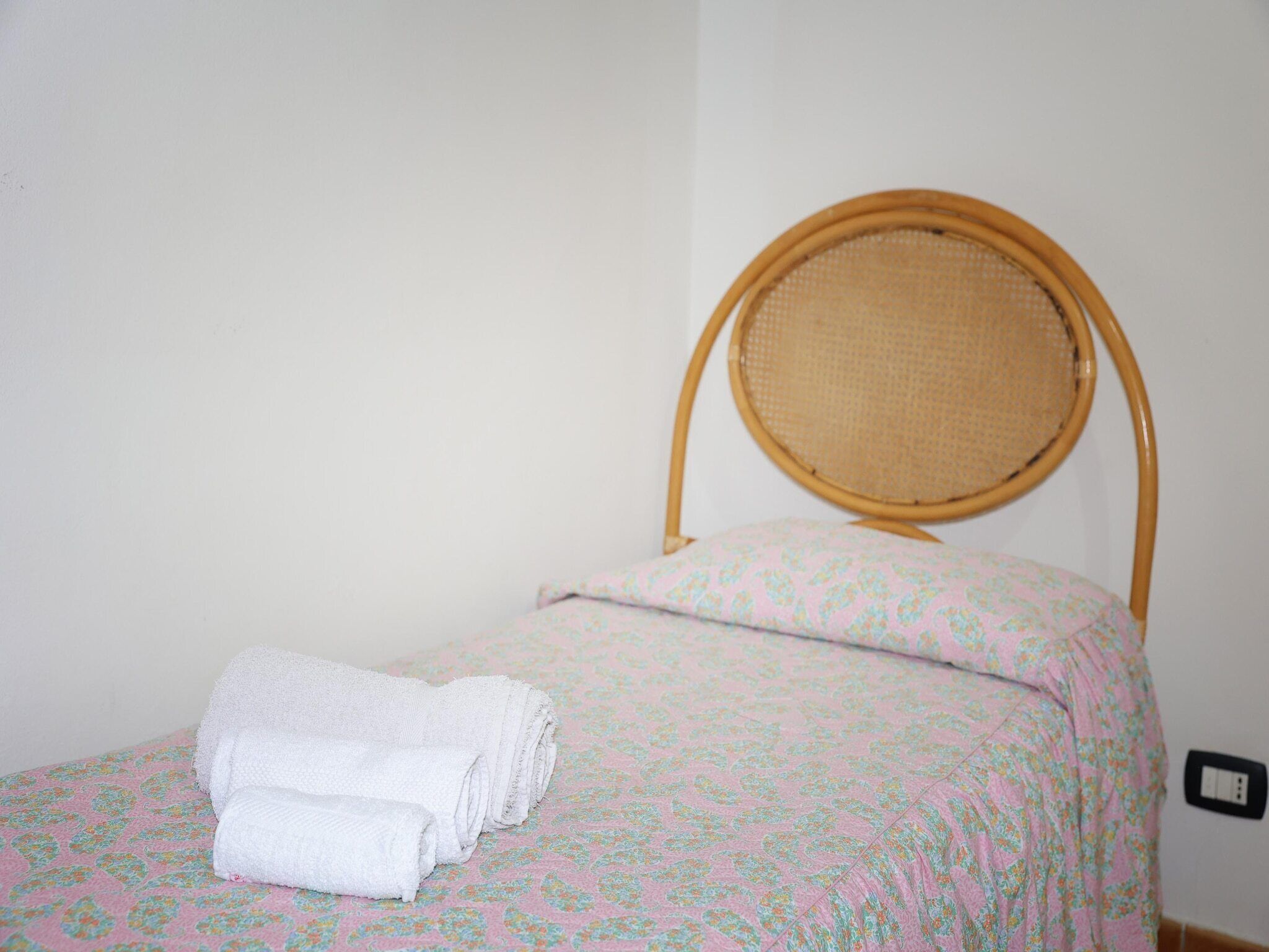 Un lit simple avec une tête de lit en osier, une couvre-lit à motifs floraux roses et deux serviettes blanches soigneusement pliées au pied du lit.