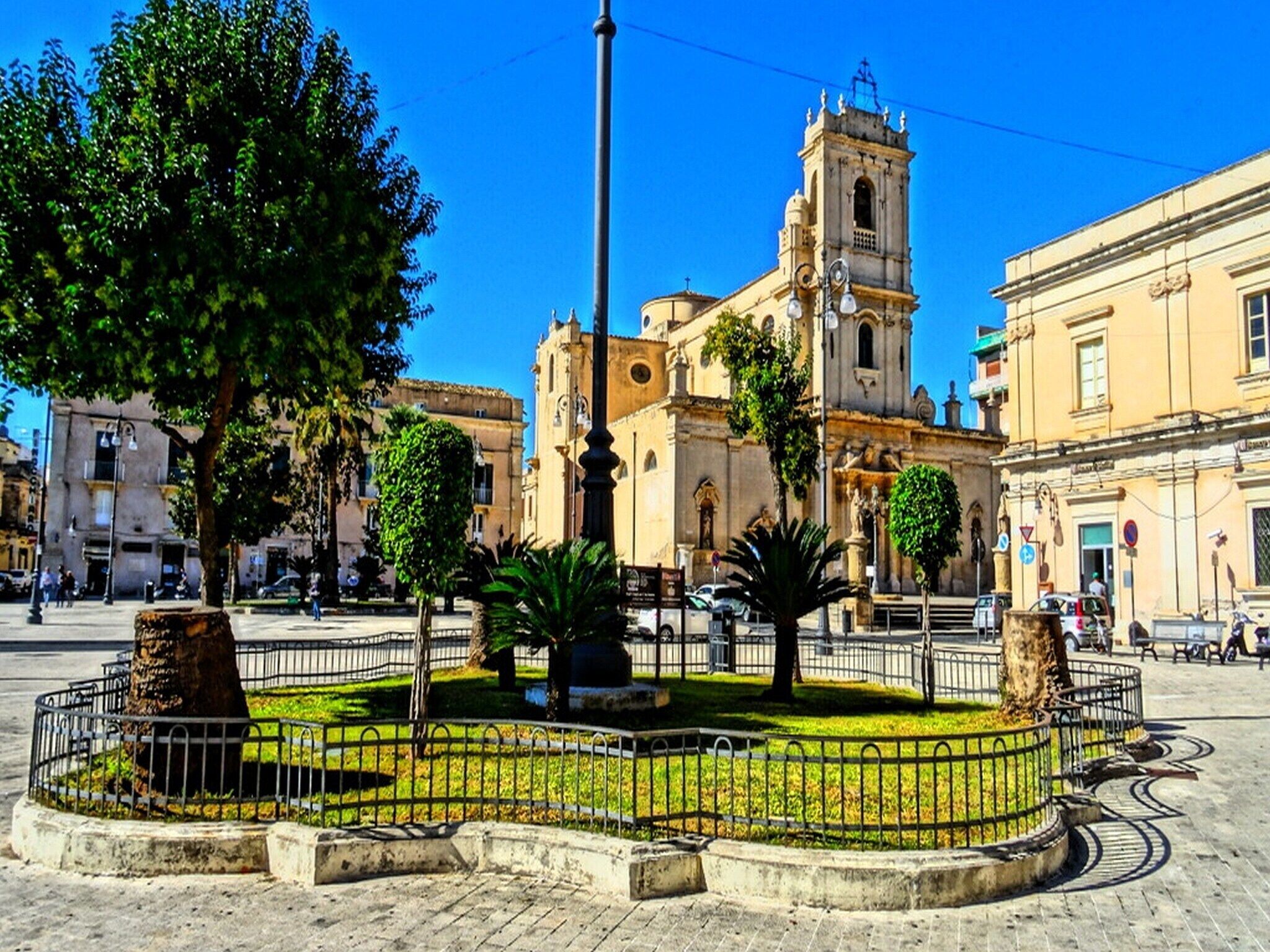 Ein historischer Platz mit einer markanten Kirche, einem zentralen Brunnen und umgebenden Bäumen.