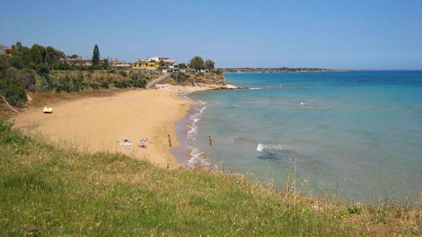 A beach with clear blue water, a sandy shore, and a few people. There are buildings in the background. - Holiday Home in Lido di Noto near Beach (Lido di Noto)