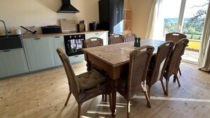 Een eetruimte met een houten tafel en rieten stoelen, een keuken met lichtblauwe kasten, en uitzicht door een groot raam.