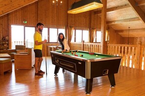 A wooden-floored room with a pool table, a person playing pool, and another person standing nearby.
