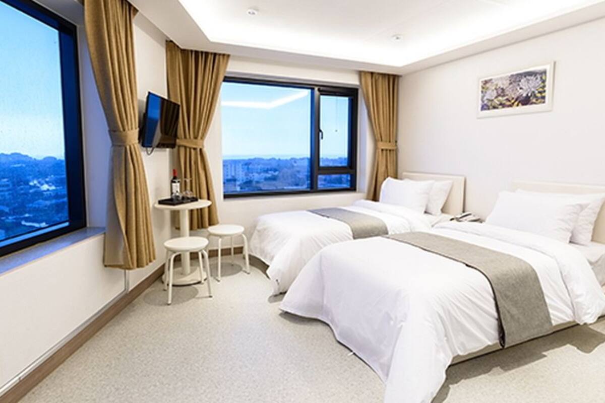 Ein Hotelzimmer mit zwei Betten, einem Fernseher, einem kleinen Tisch und einem Fenster mit Blick auf die Stadt.