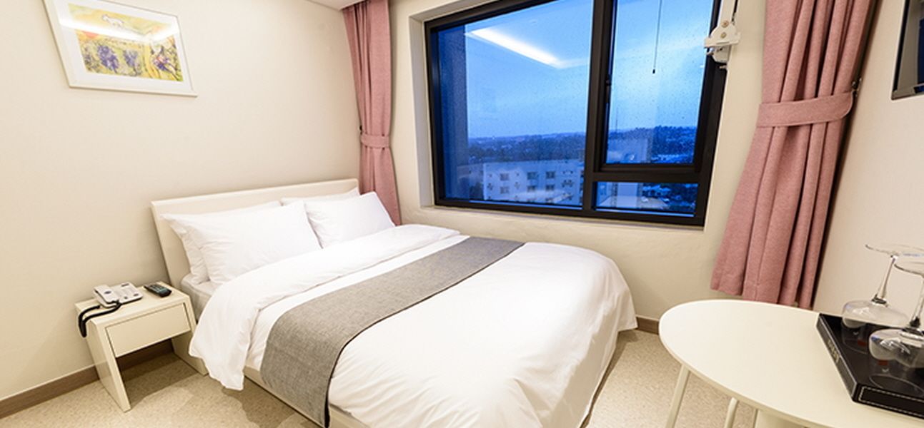 Hotellihuone, jossa on sänky, yöpöytä ja ikkuna, josta on näkymä rakennuksiin.