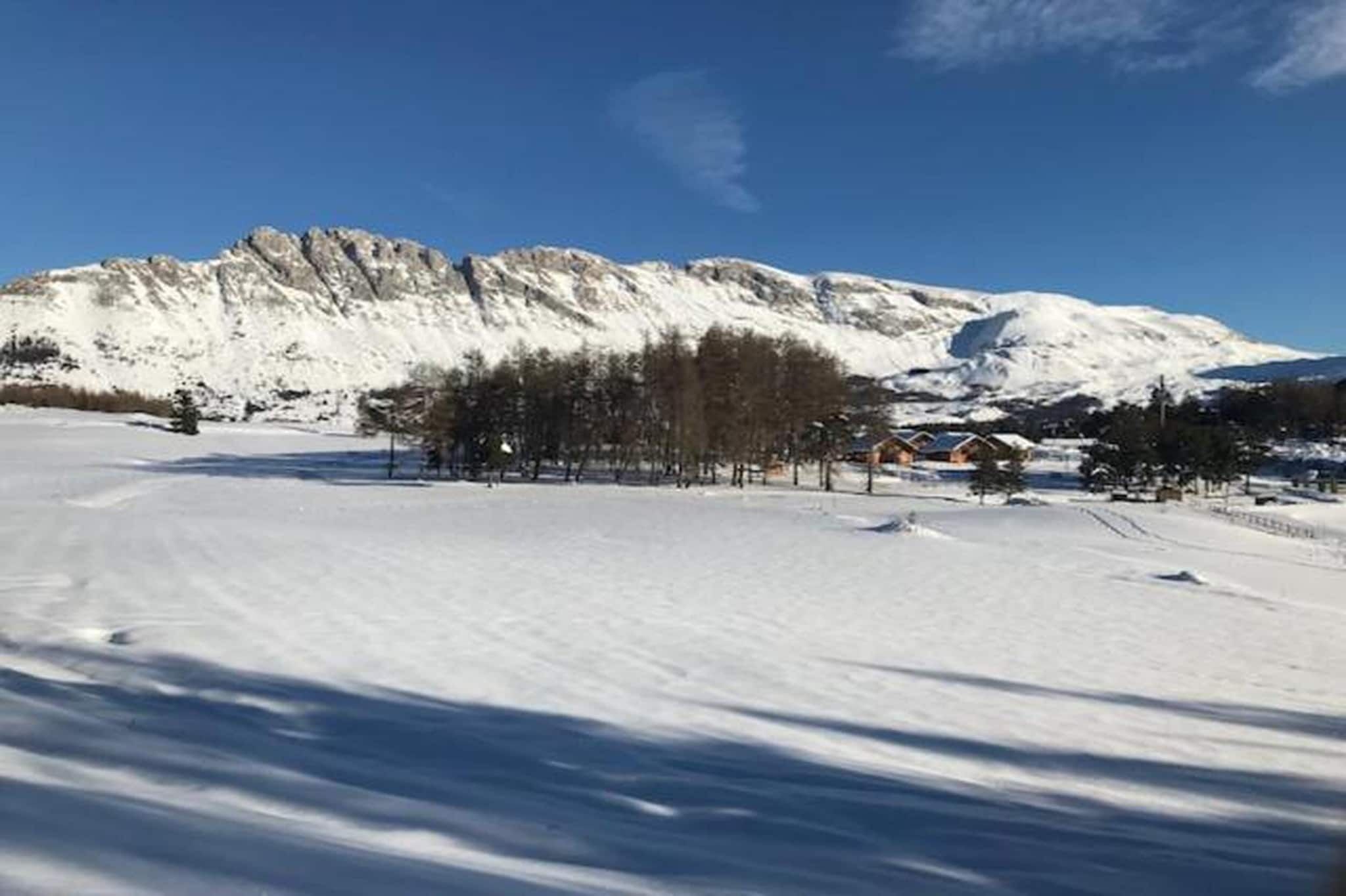 A snow-covered landscape with a mountain range, a cluster of trees, and a few buildings in the distance.