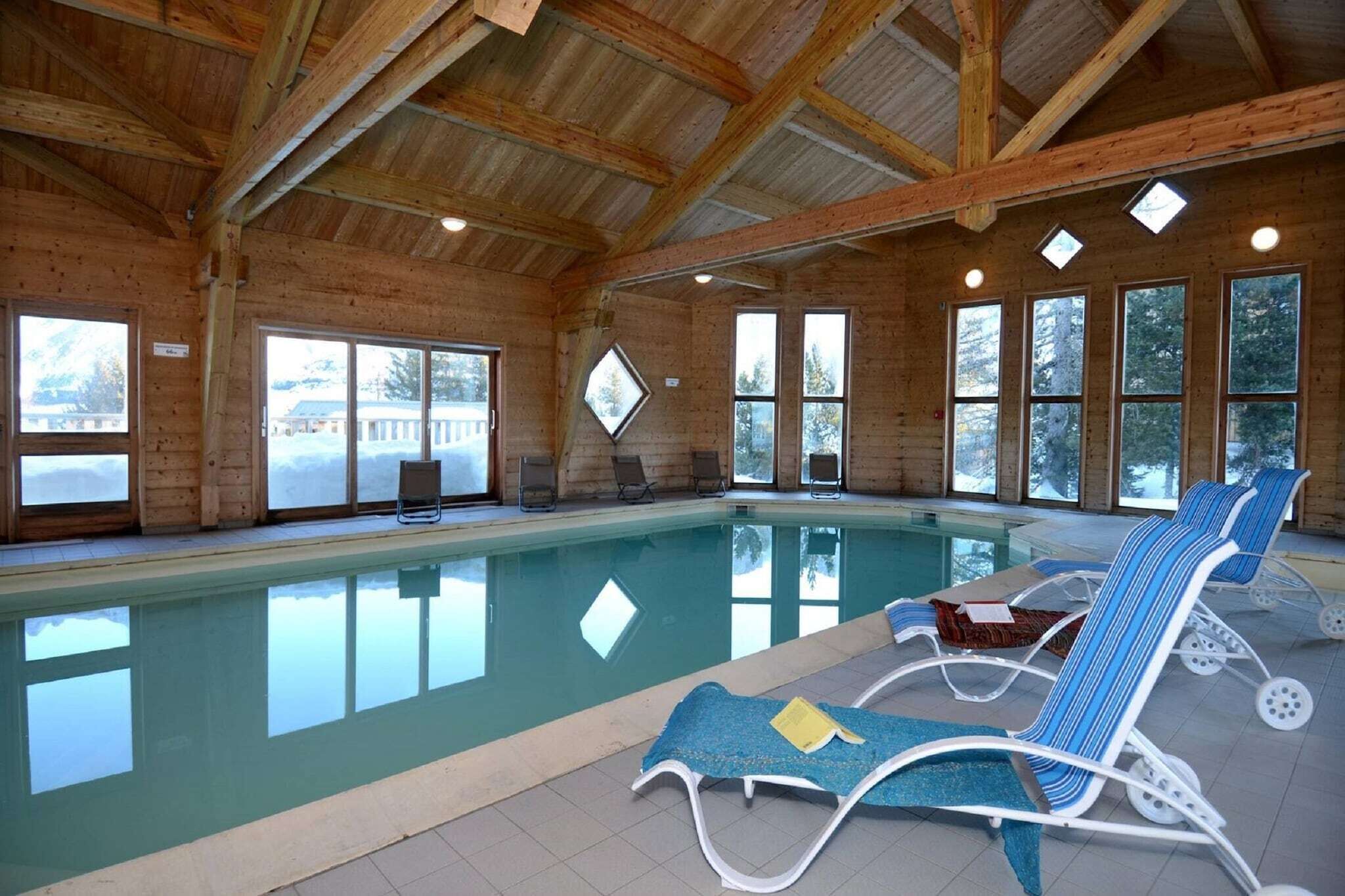 Piscina interior com paredes de vidro transparente, vigas de madeira e espreguiçadeiras.
