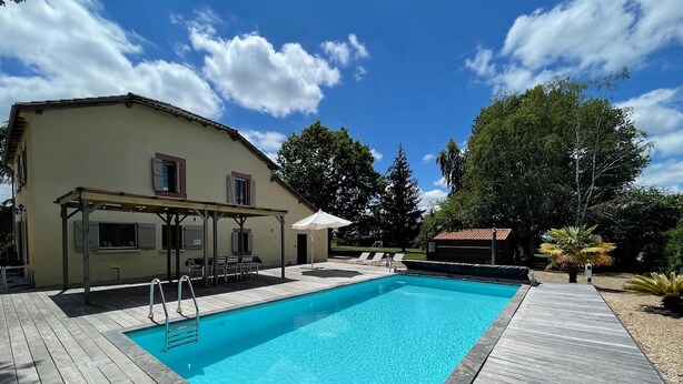 Une piscine avec une terrasse en bois, un espace salon couvert et un bâtiment doté de fenêtres et de volets.