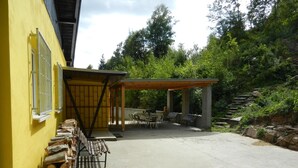 A covered outdoor seating area with tables and chairs, a stack of firewood, and a yellow building with barred windows. - Child-friendly holiday home in Moravia with a beautiful location and view (Nedvedice)