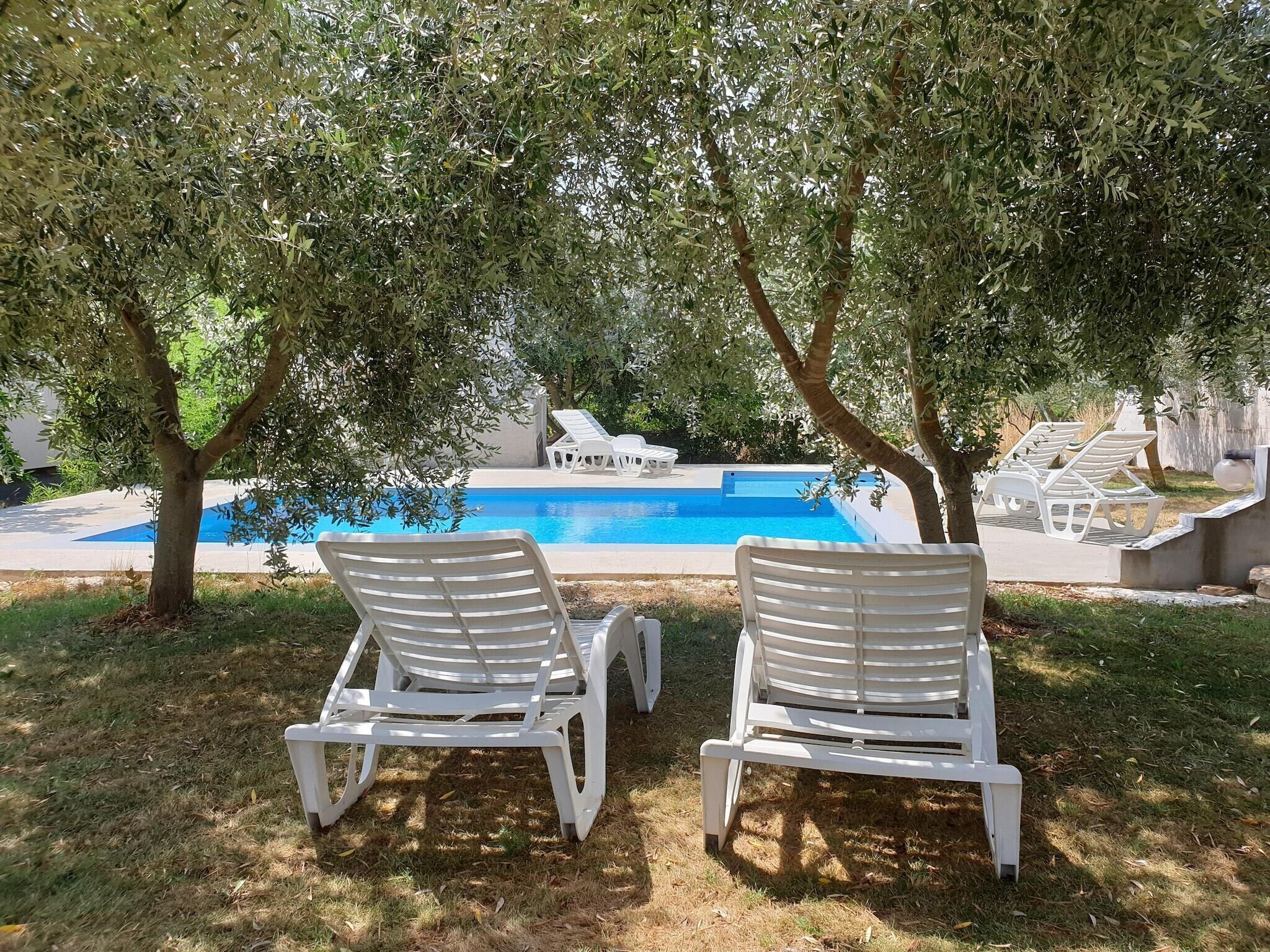 Two white lounge chairs facing a swimming pool, surrounded by trees.