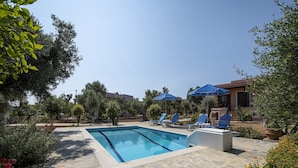 A swimming pool with stone tiles, surrounded by lounge chairs and umbrellas, with a house and trees in the background.