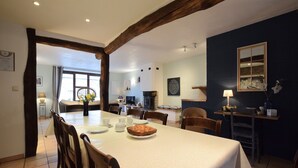A dining area with a long table, chairs, and a bread basket. There is a fireplace, a TV, and a window with curtains. - Former farmhouse in a quiet village (Manhay-Freyneux)