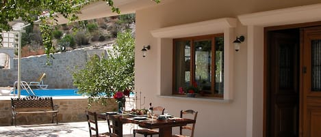 Um pátio externo com mesa e cadeiras de madeira, uma parede de pedra e uma piscina ao fundo.