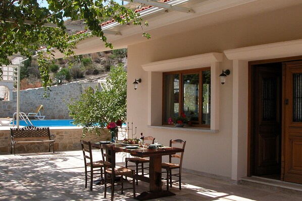An outdoor patio with a wooden table and chairs, a stone wall, and a pool in the background.