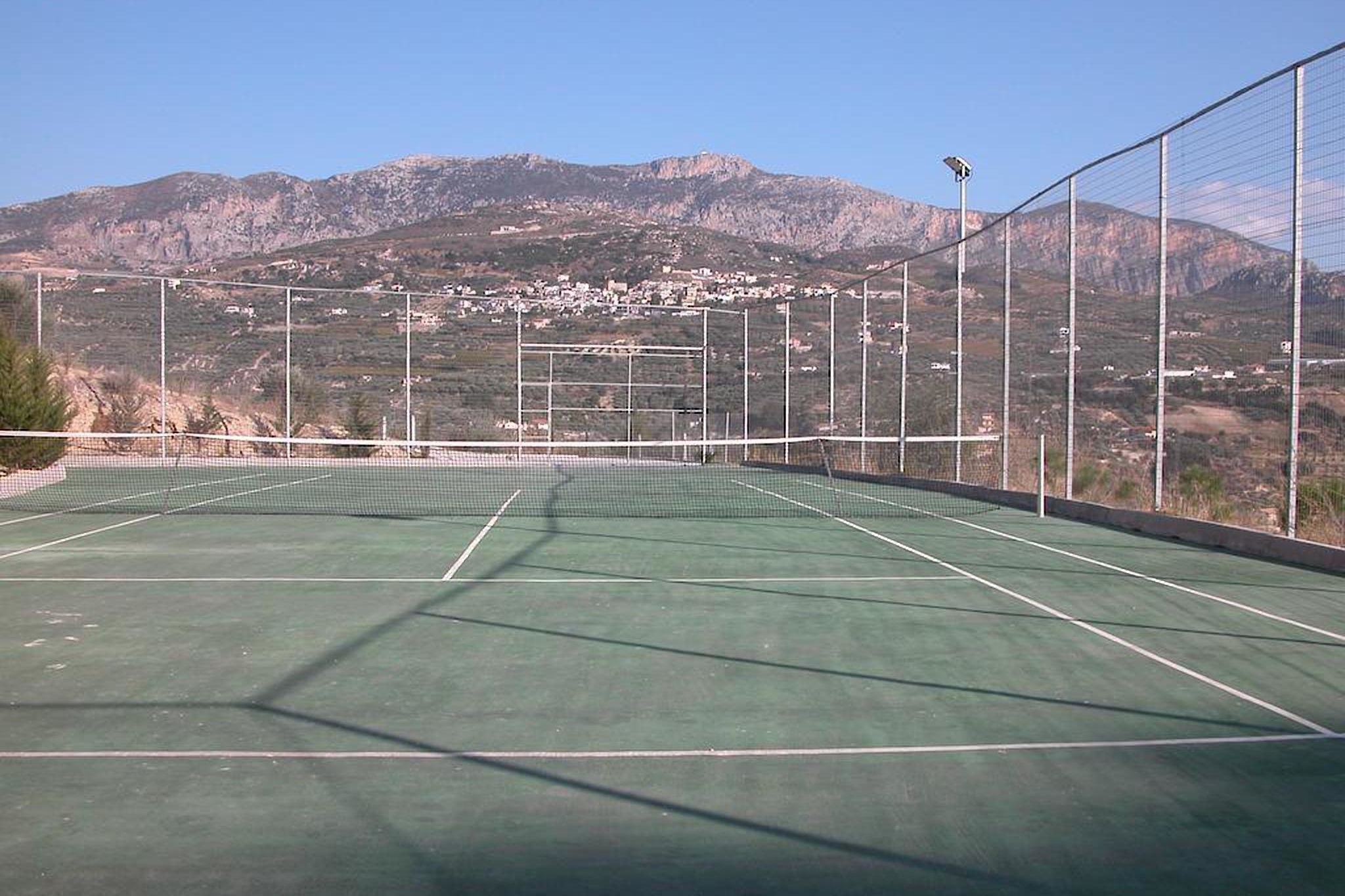 An outdoor tennis court with a net, surrounded by a fence, and a view of a town and mountains in the background.