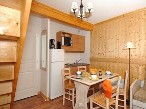 Eine kleine Küche mit Kühlschrank, Mikrowelle und Holzschränken. Es ist ein Esstisch mit Stühlen und eine kleine Tischlampe vorhanden.