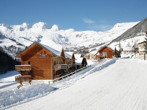 A wooden chalet with a balcony, a ski lift in the background, and snow-covered mountains.