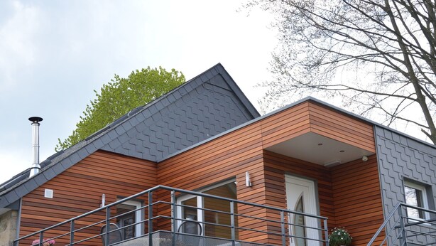 Ett modernt hus i två våningar med en grund av sten, träpanel på fasaden och ett räcke av metall på balkongen.
