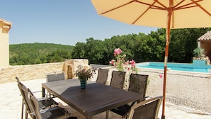 An outdoor patio with a table and chairs, a large umbrella, and a pool in the background. - Peaceful Villa in Calamane with Private Pool (Calamane)
