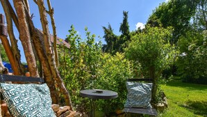 A garden patio with a wooden bench and a small round table, surrounded by lush greenery and a stone wall.