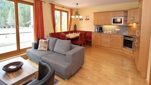 A modern living room with a grey sofa, wooden dining table, and a kitchen area with wooden cabinets and appliances. - Chalet in Saalbach near Ski Lift (Saalbach-Hinterglemm)