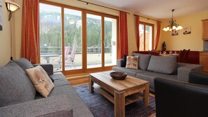 A spacious living room with a grey sofa set, a wooden coffee table, and a large sliding glass door leading to a balcony with a view of trees and mountains. - Chalet in Saalbach near Ski Lift (Saalbach-Hinterglemm)