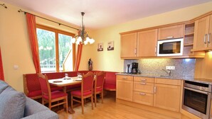 A cozy dining area with a wooden table and chairs, a kitchen with a microwave and oven, and a window with curtains. - Chalet in Saalbach near Ski Lift (Saalbach-Hinterglemm)
