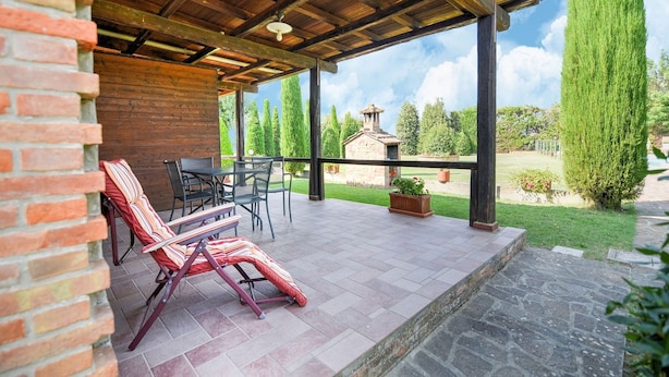 A covered patio area with a brick wall, a stone floor, and a view of a garden with a stone building and tall trees.