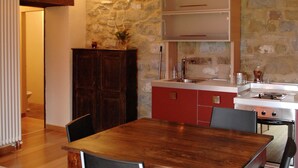 Une cuisine avec une table en bois, des chaises, un meuble rouge, un mur en pierre et un poêle.