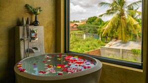A bathroom with a bathtub filled with flowers, a window with a view of greenery and a palm tree, and a towel rack with towels.