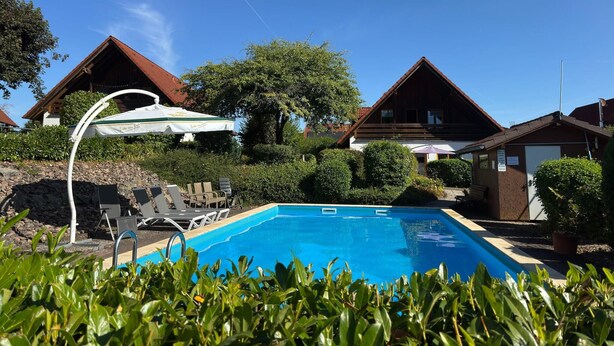 Ein Schwimmbad mit Liegestühlen und Sonnenschirmen, umgeben von Häusern mit geneigten Dächern.