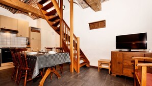 Un coin repas avec un escalier en bois, une table à manger entourée de chaises, un téléviseur posé sur un meuble en bois, et une cuisine en arrière-plan.