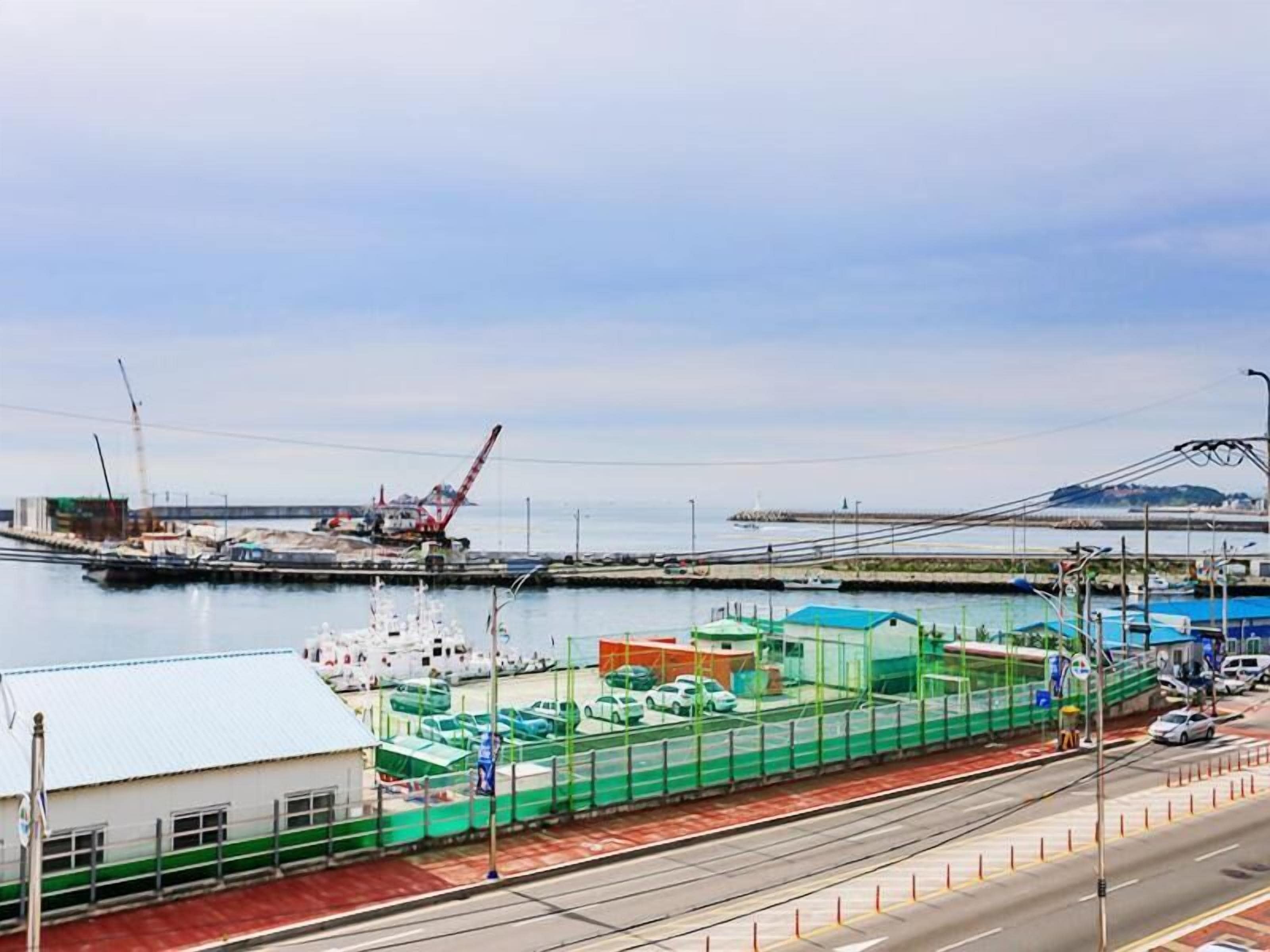 A coastal road with a green construction site, a crane, and a bridge in the background.