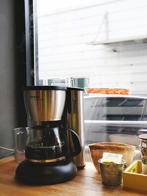 Une machine à café est posée sur une table en bois près d’une fenêtre, d’où l’on aperçoit un immeuble et une voiture à l’extérieur.