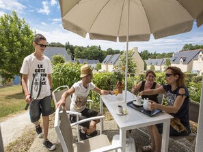 Eine Gruppe von Menschen sitzt an einem Tisch im Freien unter einem Sonnenschirm, umgeben von Grünflächen und Häusern im Hintergrund.