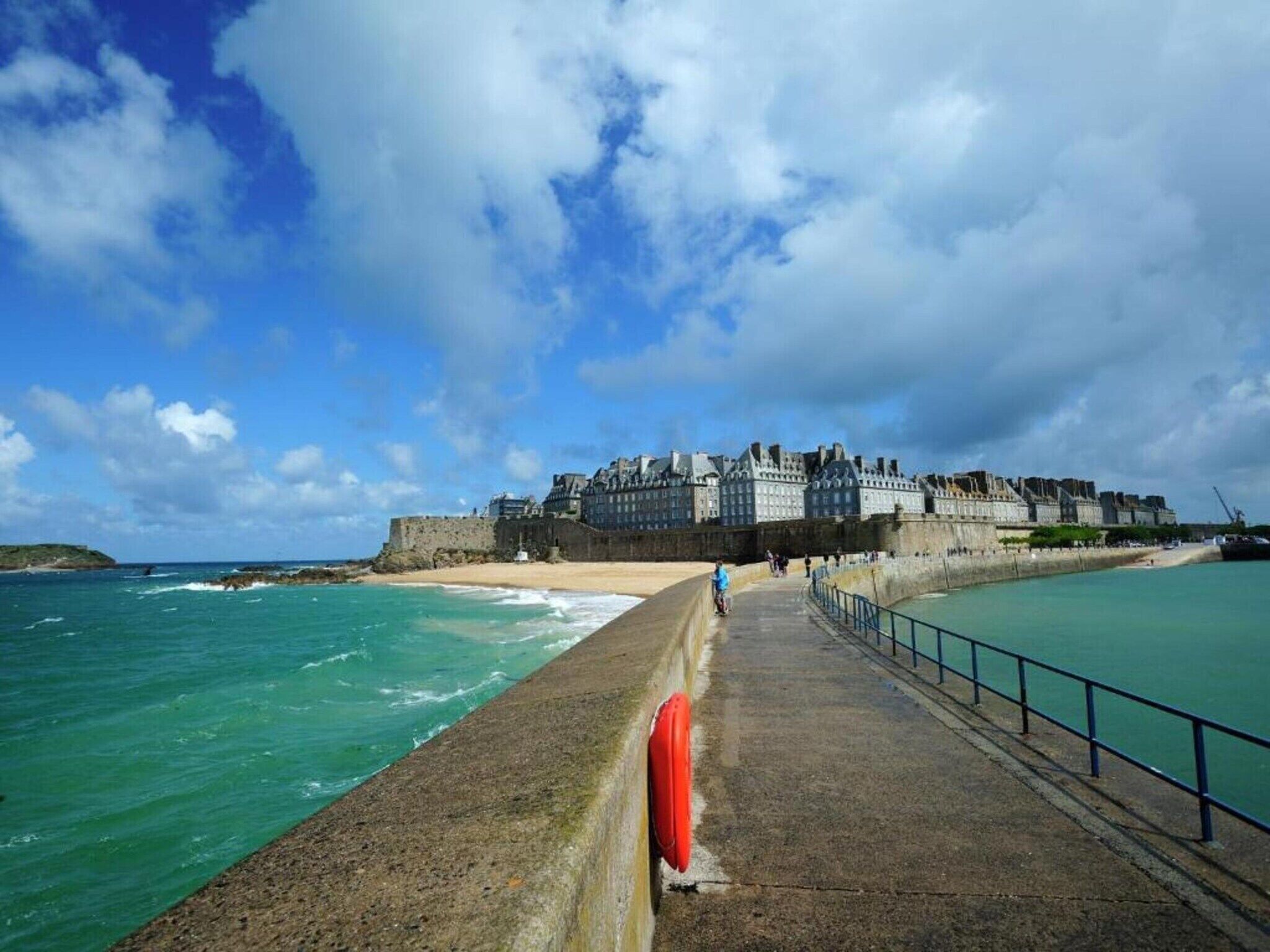 A coastal promenade with a historic building, a sandy beach, and the sea.