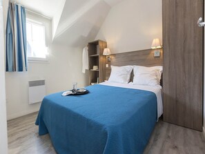 A hotel room with a bed, a bedside table with a glass and a book, a window with curtains, a wall-mounted shelf with a hat, and a wooden headboard.