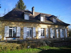 Een stenen huis met een puntige dakschild en meerdere ramen met luiken, met een bank op de voorgrond.