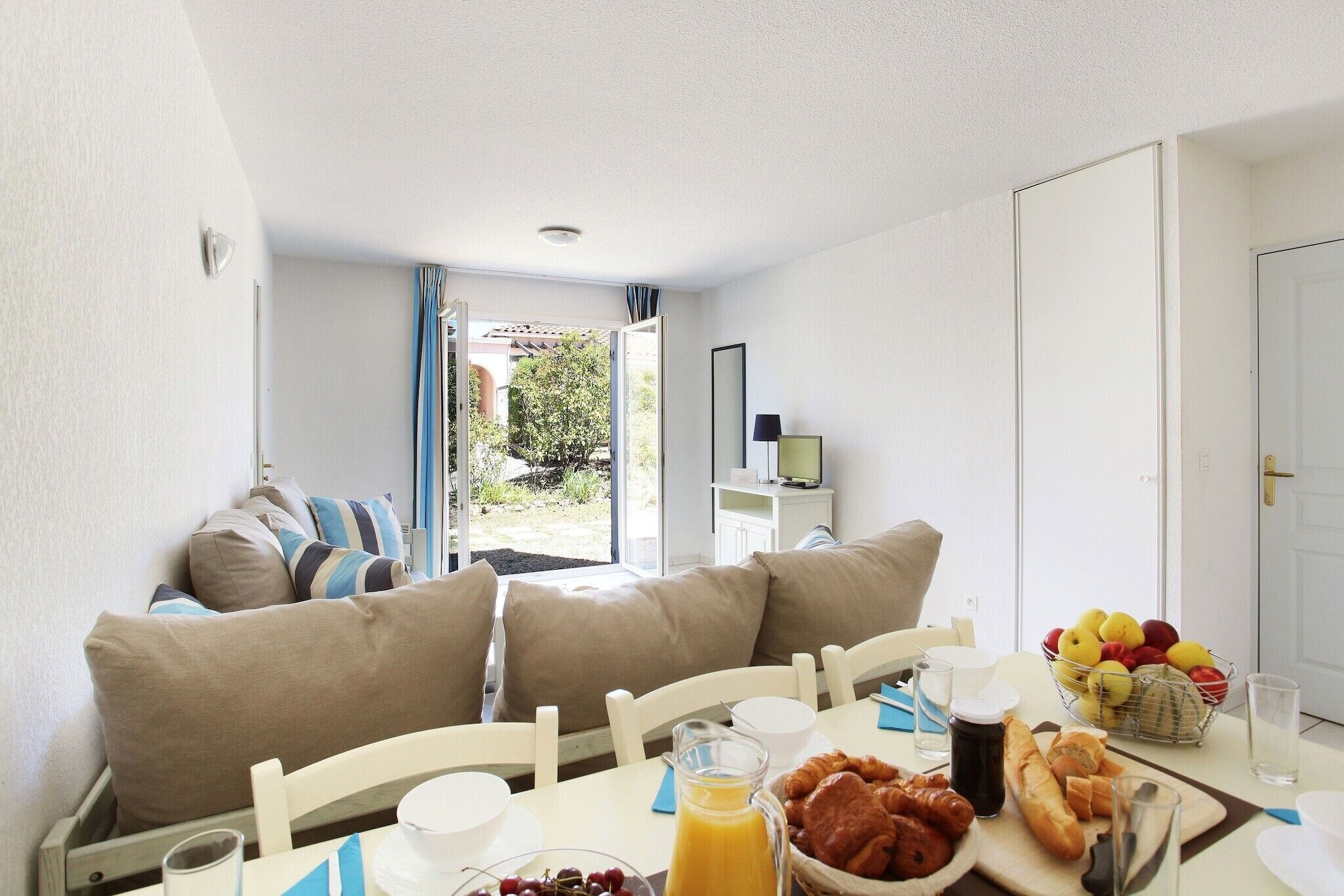 A room with a dining table set for breakfast, a sofa with cushions, a window with curtains, and a door.