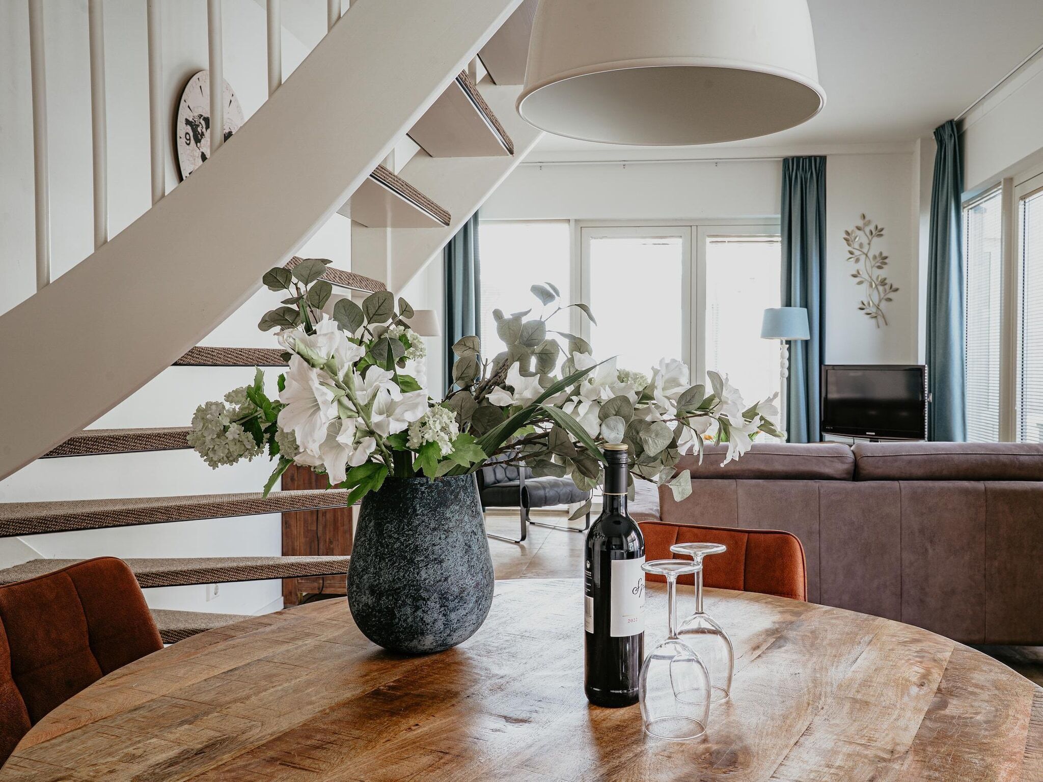Un coin repas avec une table ronde en bois, un vase rempli de fleurs et une bouteille de vin.