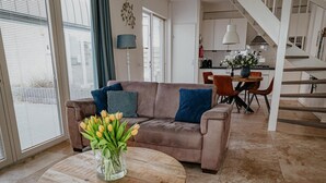 A modern living room with a brown leather sofa, a round wooden coffee table with a vase of tulips, and a dining area in the background. - Apartment in Egmond with Stunning Sea Views (Egmond aan zee)