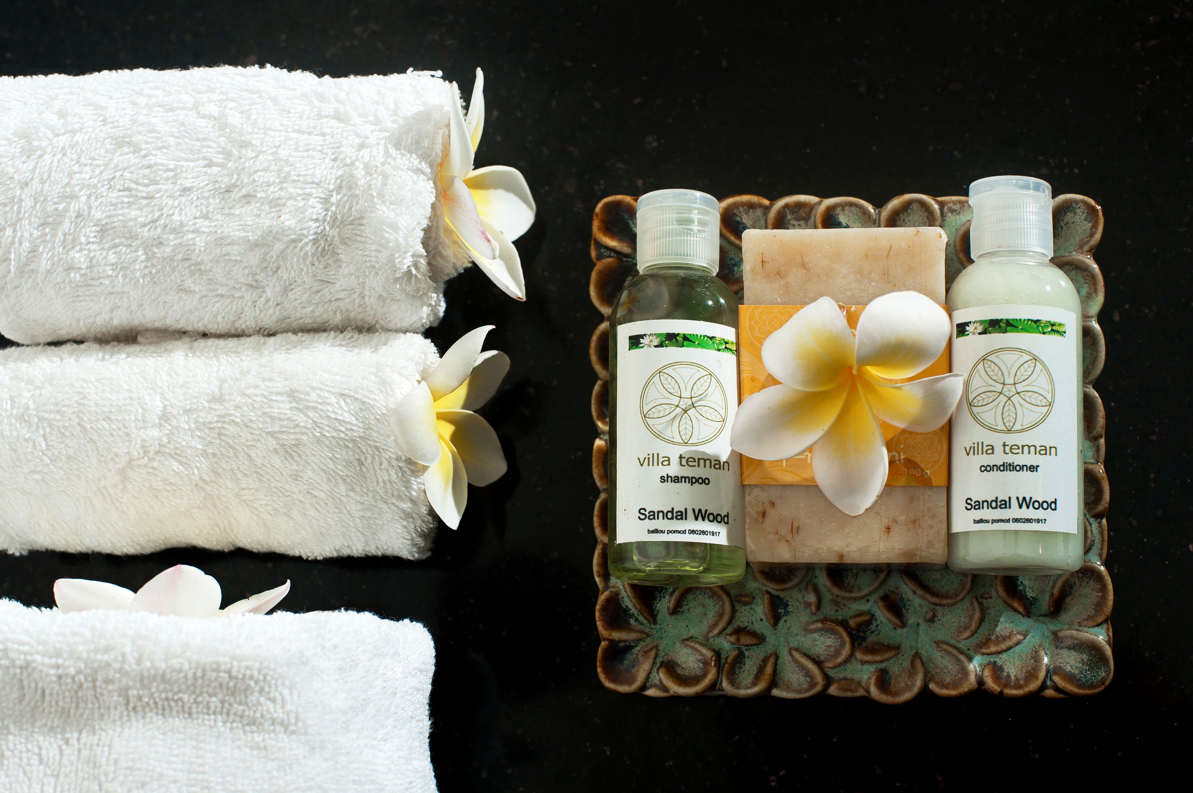 Three white towels, two bottles of 'villa teman shampoo' and 'villa teman conditioner', and a bar of soap on a decorative tray.
