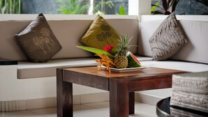 A wooden table with a fruit arrangement, a white sofa with patterned cushions, and a view of greenery through a window. - Villa Teman (Buleleng)