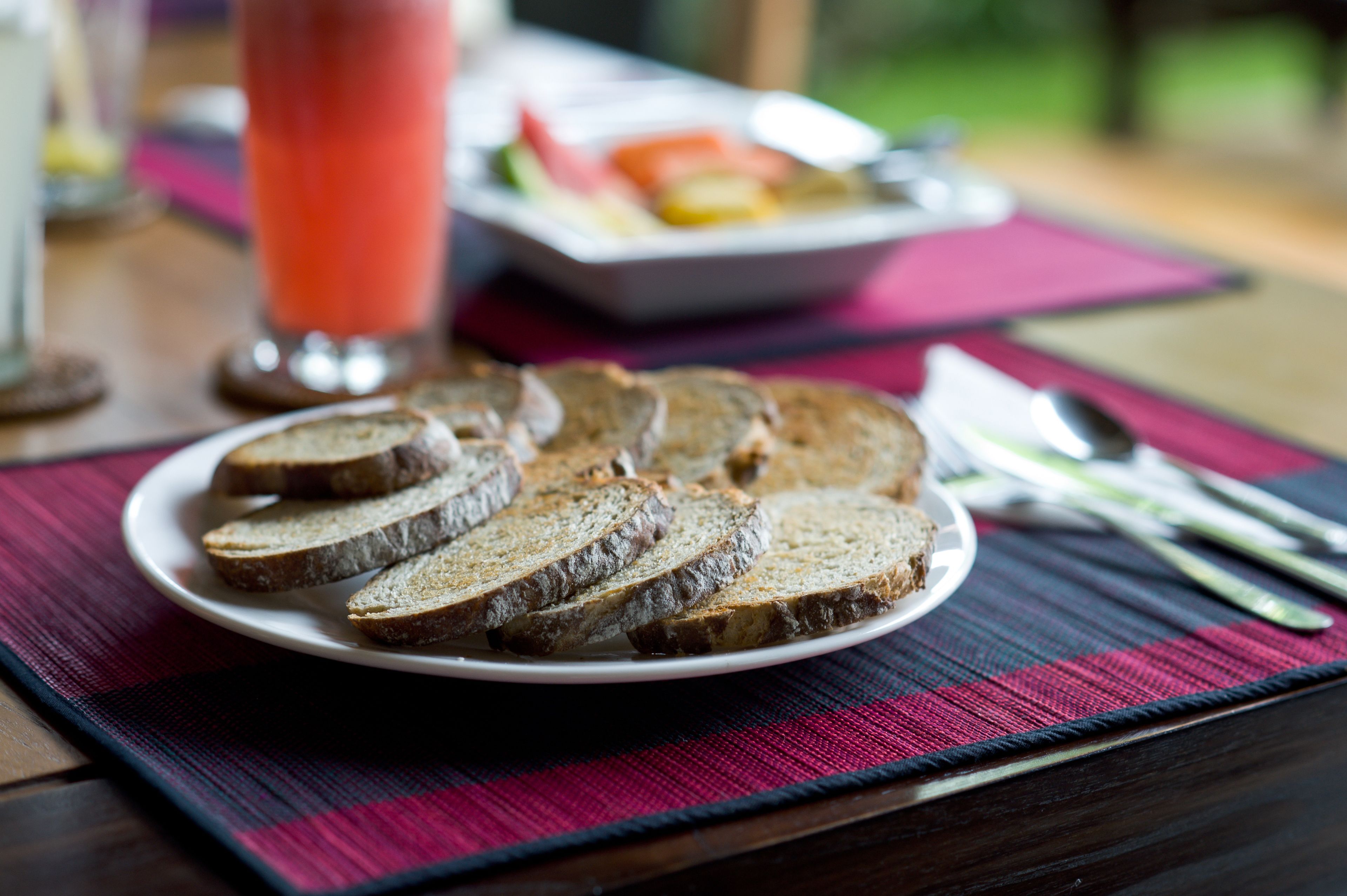 A plate of sliced bread on a placemat with a glass of red drink and a plate of fruit in the background.