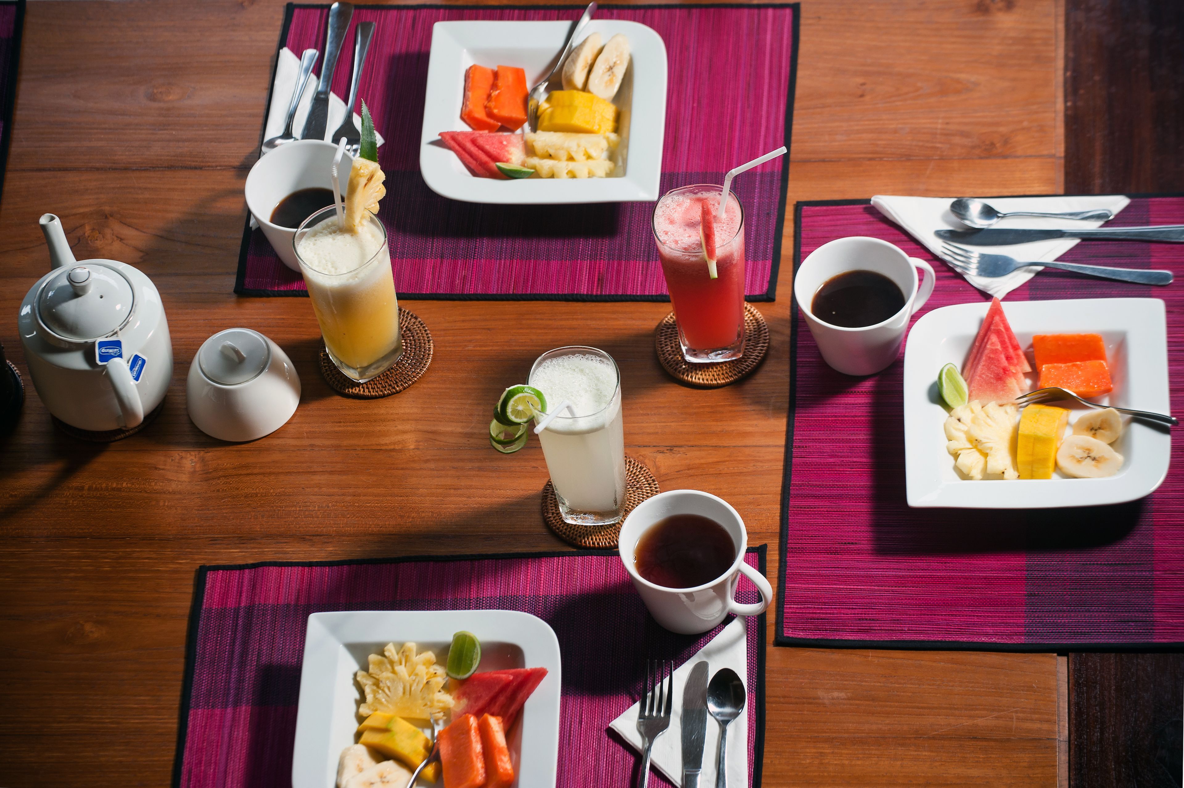 A breakfast setting with multiple plates of fruit, two cups of coffee, and a teapot on a wooden table.